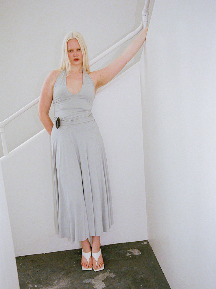 Woman in a grey dress standing in a minimalistic room with white walls and a concrete floor.